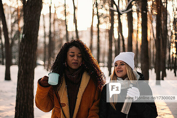 Girlfriends drinking tea and talking while walking in park in winter