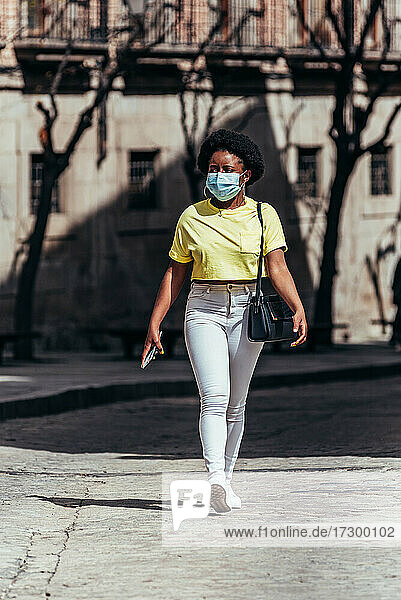 Schwarzes afroamerikanisches Mädchen mit Gesichtsmaske  das eine Stadtstraße entlanggeht.