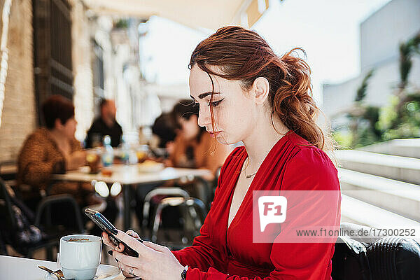 Junge rothaarige Frau in roter Bluse  die auf einer Terrasse Kaffee trinkt und dabei ihr Mobiltelefon benutzt.
