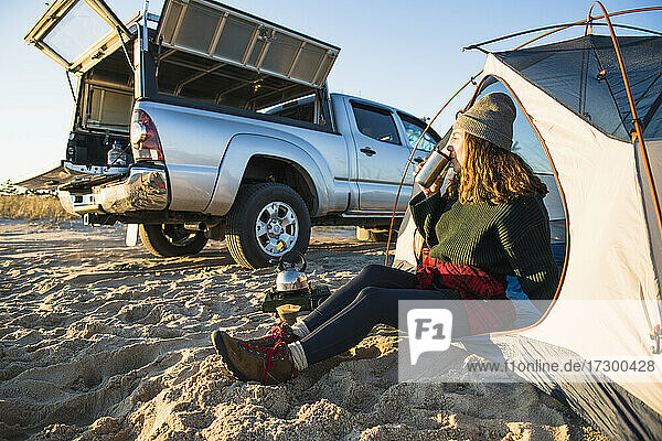 Junge Frau genießt Kaffee im Reisebecher beim Camping am Strand