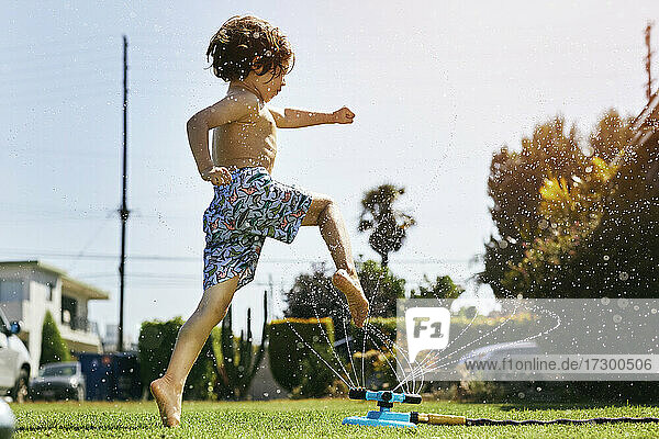Shirtless boy jumping over sprinkler in backyard against sky
