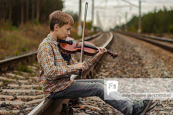 Ein hübscher blonder Junge sitzt auf den Schienen der Eisenbahn und spielt die v