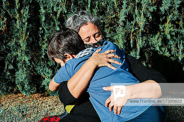 Nahaufnahme einer glücklichen Mutter mit einer Maske  die ihren Sohn umarmt  während sie im Park im Gras sitzt. Neue Normalität