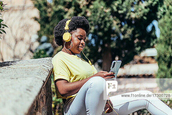 Schwarzes afroamerikanisches Mädchen sitzt und entspannt  während sie mit ihrem Handy und Kopfhörern Musik hört.