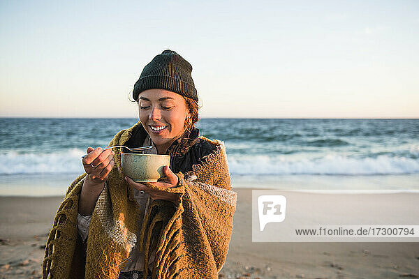 Junge Frau genießt eine Schüssel Suppe beim Camping mit dem Auto am Strand im Herbst