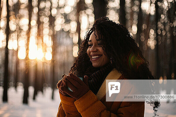 Happy young woman holding cup of tea standing in park during winter