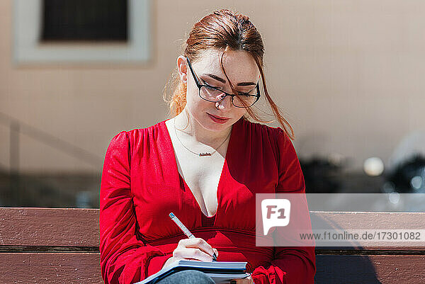 Junge rothaarige Frau in roter Bluse  die auf einer Bank sitzt und etwas in ihr Notizbuch schreibt.