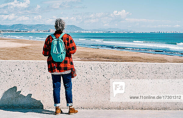 Frau mittleren Alters von hinten mit Rucksack  die an einem sonnigen Tag auf das Meer schaut