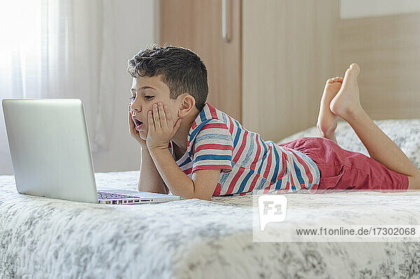 Junger Junge  der auf dem Bett liegt und einen Laptop benutzt. Online-Klasse Lektion