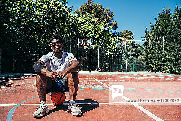 Porträt eines schwarzen Afro-Jungen  der auf seinem Ball auf dem Basketballplatz sitzt.