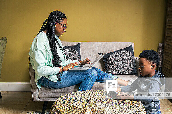 Mutter benutzt Laptop  während Sohn ein Spiel auf dem Handy im Wohnzimmer zu Hause spielt
