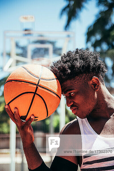 Porträt eines schwarzen afroamerikanischen Jungen  der auf einem städtischen Basketballplatz einen Basketball in die Stirn hält.