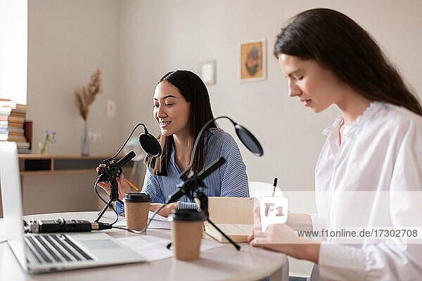 Asiatische Frau spricht in ein Mikrofon neben einem Kollegen  der während eines Podcasts Notizen macht