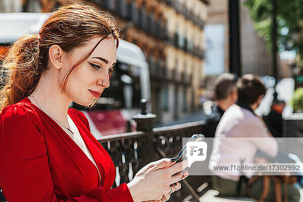 Young red-haired girl dressed in a red blouse using her mobile phone in a city square.
