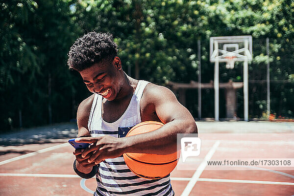 Porträt eines schwarzen afroamerikanischen Jungen  der mit seinem Mobiltelefon auf einem städtischen Basketballplatz eine Nachricht sendet.