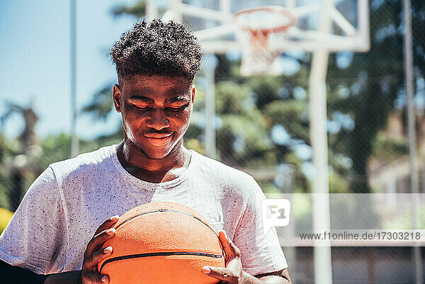Porträt eines schwarzen afroamerikanischen Jungen  der auf einem städtischen Basketballplatz einen Basketball gegen seine Brust hält.