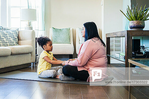 Grandmother practicing meditation with granddaughter while sitting on exercise mat in living room at home