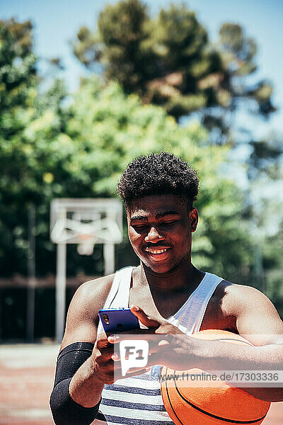 Porträt eines schwarzen afroamerikanischen Jungen  der sein Mobiltelefon auf einem städtischen Basketballplatz benutzt.