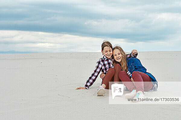 The sisters are hugging while sitting on the sandy slope.