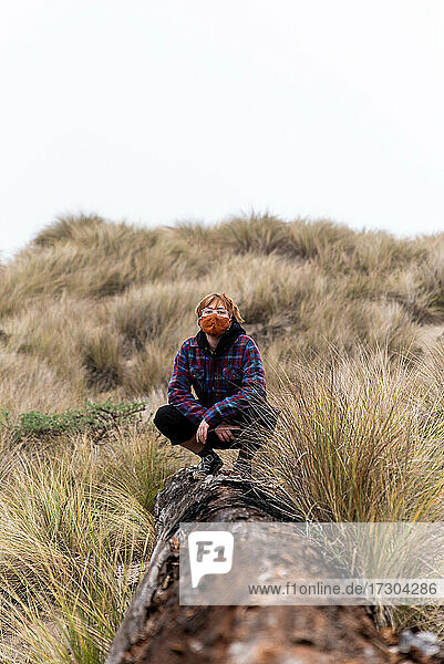 Teen with mask sitting on logs at coastal sand dune on foggy day