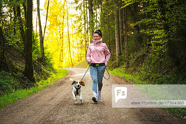 Young woman and dog running together in sunny forest.
