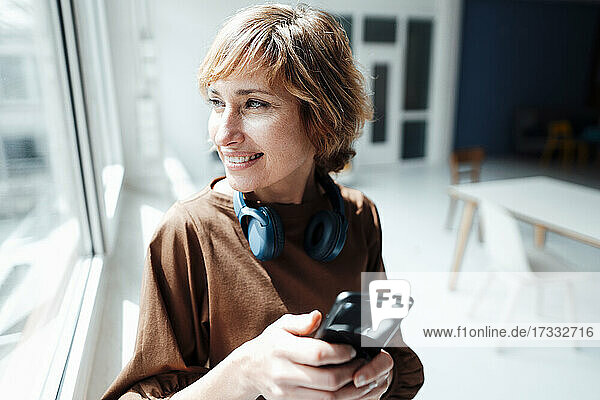 Lächelnde Geschäftsfrau mit Kopfhörern und Smartphone schaut durch ein Fenster im Büro