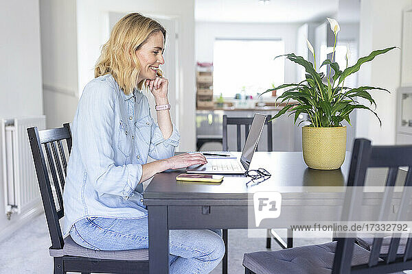 Frau mit Hand am Kinn  die einen Laptop benutzt  während sie zu Hause auf einem Stuhl sitzt