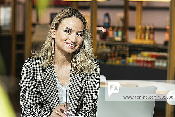 Smiling beautiful businesswoman holding pen while sitting with laptop at coffee shop