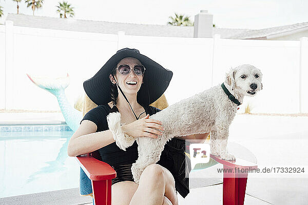 Woman in hat smiling while sitting on chair with dog at patio during vacation