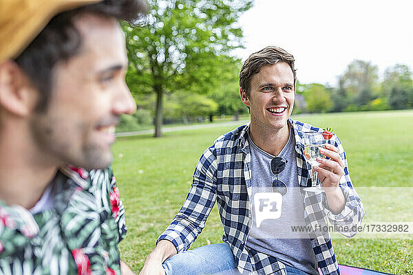 Smiling man holding cocktail while sitting by friend at park