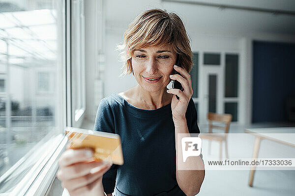 Geschäftsfrau  die eine Kreditkarte in der Hand hält  während sie mit einem Smartphone im Büro telefoniert