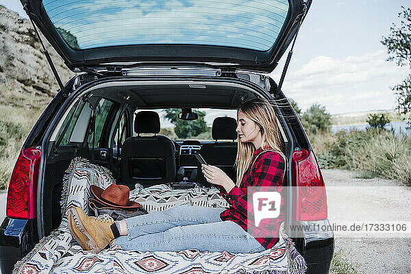 Junge Frau sendet Textnachrichten über ihr Smartphone  während sie im Kofferraum eines Autos sitzt