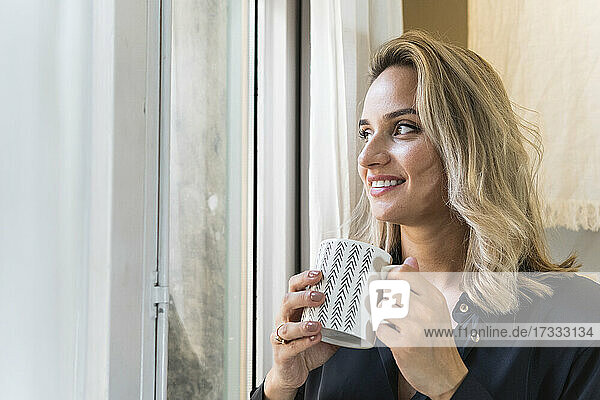 Nachdenkliche Geschäftsfrau  die eine Tasse in der Hand hält  während sie durch das Fenster ihres Hauses schaut