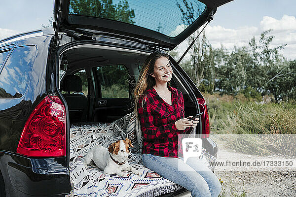Glückliche Frau  die ein Mobiltelefon hält  sitzt neben einem Hund im Kofferraum eines Autos