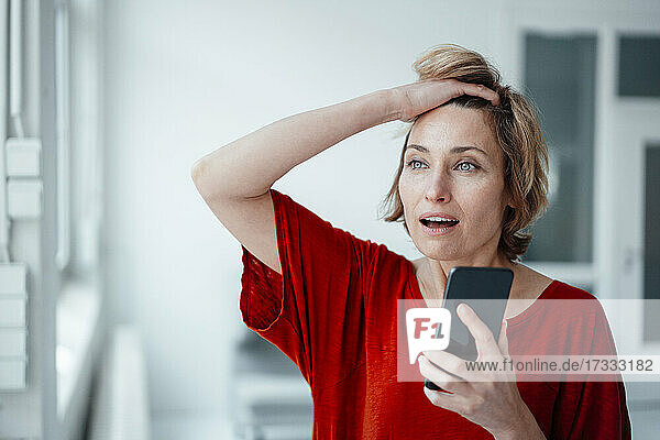 Geschäftsfrau mit offenem Mund und Hand in den Haaren hält Smartphone im Büro