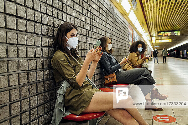 Frau mit Gesichtsmaske  die ein Mobiltelefon benutzt  während sie in sozialer Distanz in der U-Bahn sitzt