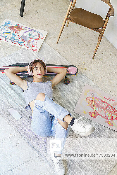 Junge Malerin mit Händen hinter dem Kopf entspannt auf einem Skateboard im Atelier