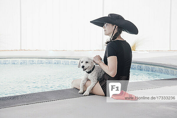 Frau mit Hut schaut weg  während sie mit ihrem Hund am Pool im Innenhof sitzt