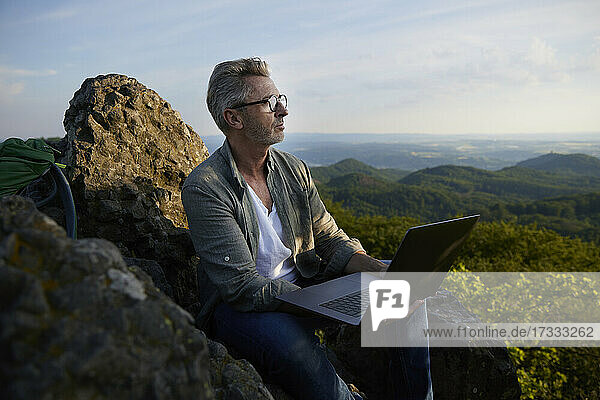 Mann hält Laptop und sitzt auf einem Berg