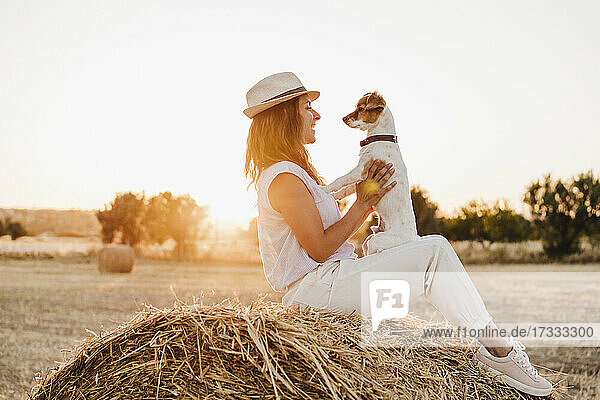 Woman wearing hat playing with dog on strawb bale