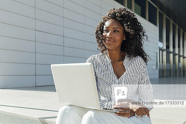 Nachdenkliche Geschäftsfrau  die mit ihrem Laptop vor einem Bürogebäude sitzt und wegschaut