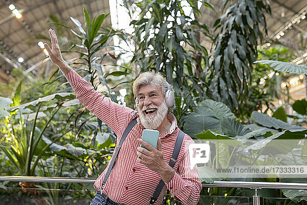 Fröhlicher reifer Mann mit erhobener Hand tanzt beim Hören von Musik vor Pflanzen