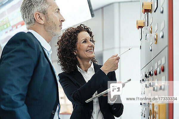 Geschäftsfrau mit digitalem Tablet  die auf eine Maschine zeigt  während sie neben einem Kollegen im Kontrollraum steht