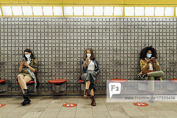 Freunde mit Schutzmaske telefonieren mit dem Handy  während sie in sozialer Distanz in der U-Bahn sitzen