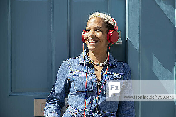 Lächelnde Frau hört Musik über Kopfhörer vor einer blauen Tür
