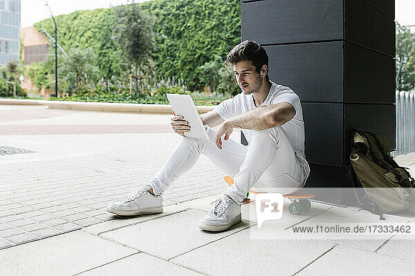 Young man using digital tablet on skateboard at column