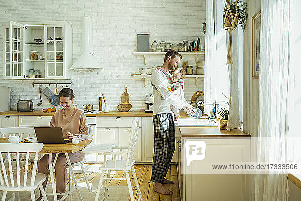 Lächelnde Geschäftsfrau mit Laptop und Mann mit Tochter beim Reinigen des Tellers in der Küche