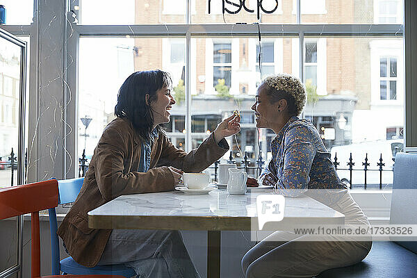 Glückliche junge Frau füttert ihre Freundin im Café sitzend mit Essen