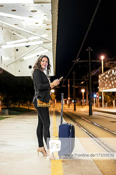 Lächelnde weibliche Fachkraft mit Gepäck  die ihr Handy in der Hand hält  während sie auf eine nächtliche Straßenbahnstation wartet