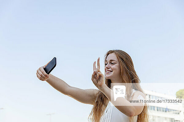 Lächelnde Geschäftsfrau gestikuliert  während sie ein Selfie mit ihrem Mobiltelefon vor dem Himmel macht